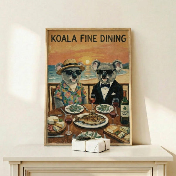 May include: Framed vintage-style poster showing two koalas in sunglasses dining at a waterfront restaurant table. One wears a tropical shirt and hat, the other a black tuxedo. The sunset beach scene features wine glasses, fish dinner, eucalyptus leaves, and cheese. Displayed on light wood shelf with small wrapped gift box beside frame.