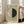 May include: Framed museum exhibition poster leaning against white wall next to beige upholstered chair with person's hands visible. Poster features cream background with large circle divided into light and dark halves, geometric black lines, and red triangle accent. Text at top and bottom in black lettering. Natural light streams through tall windows creating soft shadows on wooden floor.