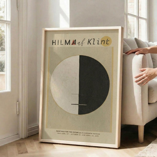 May include: Framed museum exhibition poster leaning against white wall next to beige upholstered chair with person's hands visible. Poster features cream background with large circle divided into light and dark halves, geometric black lines, and red triangle accent. Text at top and bottom in black lettering. Natural light streams through tall windows creating soft shadows on wooden floor.