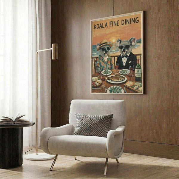 May include: Vintage-style poster showing two koalas dressed formally at a dining table with wine and food, displayed on wood-paneled wall above beige upholstered chair with geometric pillow. Modern living room setting with brass floor lamp and side table. Poster has warm sunset colors and retro travel advertisement aesthetic.
