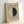 May include: Framed museum exhibition poster leaning against white wall. Features minimalist design with cream background, circular motif split between white and dark green halves, and text at top and bottom. Light wood frame complements neutral color palette. Soft natural lighting creates subtle shadows on wall.