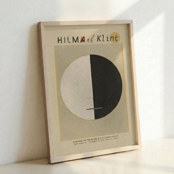 May include: Framed museum exhibition poster leaning against white wall. Features minimalist design with cream background, circular motif split between white and dark green halves, and text at top and bottom. Light wood frame complements neutral color palette. Soft natural lighting creates subtle shadows on wall.
