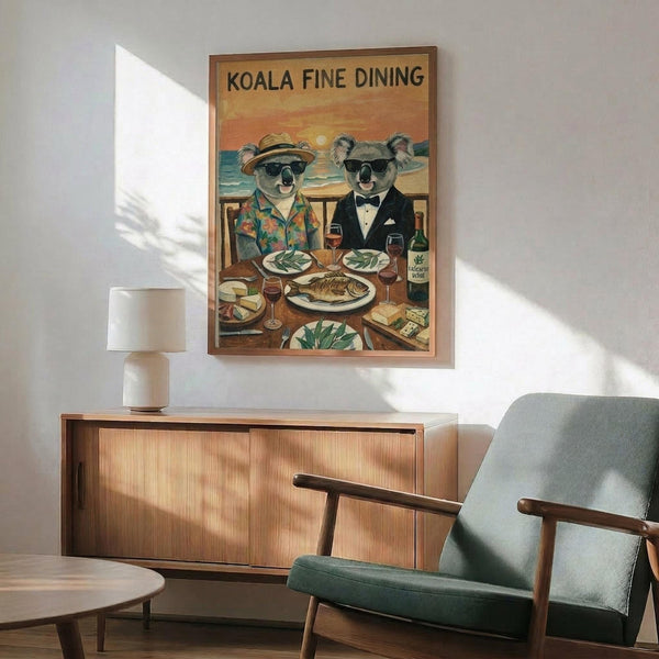 May include: Vintage-style poster showing two koalas in sunglasses dining at a wooden table with ocean sunset backdrop. One wears a Hawaiian shirt and sun hat, the other a formal tuxedo. Table displays fish, wine glasses, eucalyptus leaves, and cheese. Framed artwork hangs above mid-century modern wood credenza with white ceramic lamp. Sage green upholstered chair with wooden arms visible in foreground. Warm sunlight streams across white walls creating dramatic shadows.