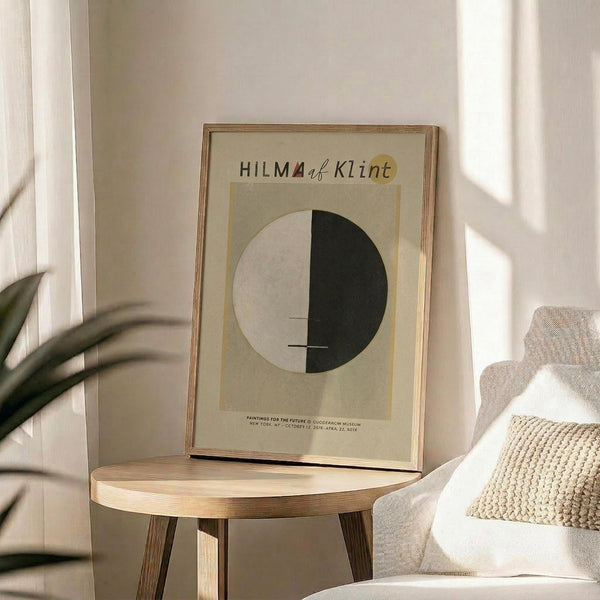 May include: Framed art print displayed on wooden round side table next to beige upholstered chair with knit throw pillow. Print features geometric design with half black, half white circle on cream background with "HILMA af Klint" text at top. Natural sunlight creates shadows across white wall. Green plant leaves visible in corner of minimalist, neutral-toned room setting.