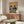 May include: Vintage-style poster featuring two koalas in sunglasses dining at a seaside restaurant, one wearing a Hawaiian shirt and hat, the other in a black tuxedo, sharing wine and fish with ocean sunset backdrop. Displayed above a mid-century modern walnut credenza with white lamp in minimalist living room setting with cream chair and glass coffee table.