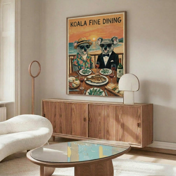 May include: Vintage-style poster featuring two koalas in sunglasses dining at a seaside restaurant, one wearing a Hawaiian shirt and hat, the other in a black tuxedo, sharing wine and fish with ocean sunset backdrop. Displayed above a mid-century modern walnut credenza with white lamp in minimalist living room setting with cream chair and glass coffee table.