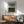 May include: Abstract art print with horizontal black lines between blue and green watercolor sections, displayed in thin wood frame above mid-century modern walnut credenza. Green upholstered armchair with wooden arms sits nearby, white ceramic table lamp and round wood coffee table complete the minimalist living room scene with natural sunlight casting shadows on white walls.