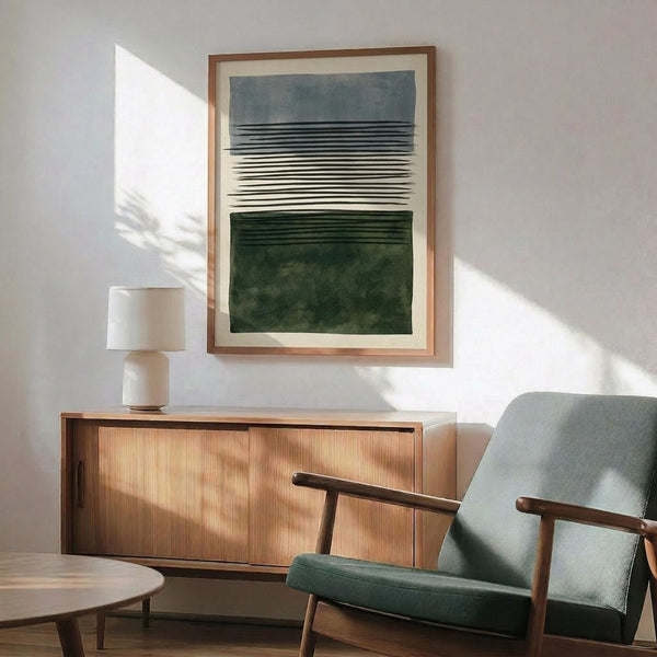 May include: Abstract art print with horizontal black lines between blue and green watercolor sections, displayed in thin wood frame above mid-century modern walnut credenza. Green upholstered armchair with wooden arms sits nearby, white ceramic table lamp and round wood coffee table complete the minimalist living room scene with natural sunlight casting shadows on white walls.
