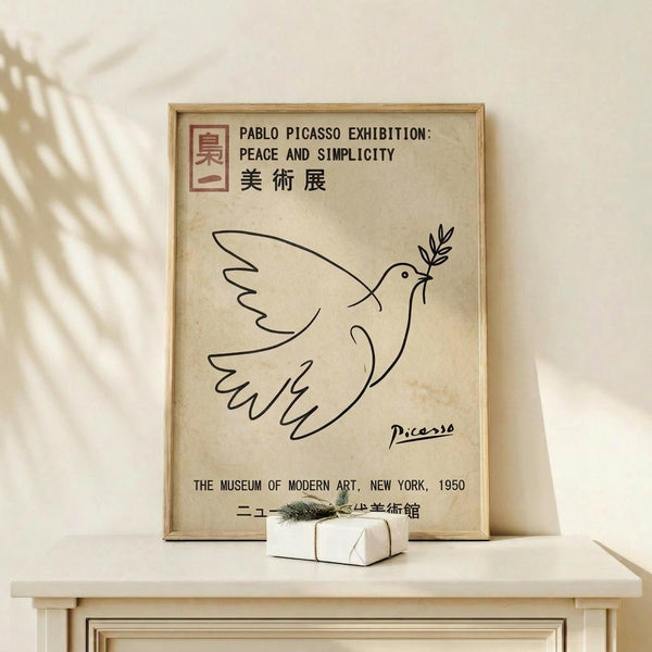 May include: Framed beige poster with black line drawing of dove carrying olive branch, titled "Pablo Picasso Exhibition: Peace and Simplicity" with Japanese characters and "The Museum of Modern Art, New York, 1950" text below. Light wood frame displayed on white wooden shelf against cream wall with plant shadows, small wrapped gift box with greenery beside frame.