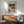 May include: Framed Paul Klee abstract geometric art print hanging above mid-century modern wooden credenza with table lamp. Green upholstered armchair with wooden frame sits beside small round side table. Colorful poster features triangular shapes, circles, and rectangles in blues, yellows, oranges, and greens against cream background. Natural sunlight creates shadows on white wall in modern living room setting.