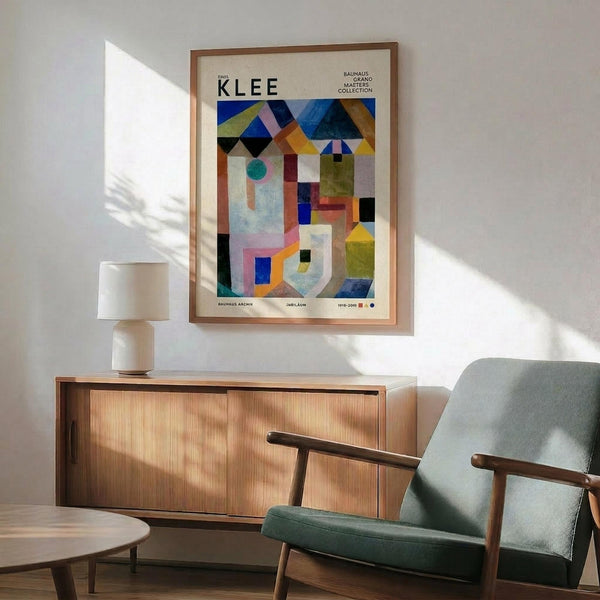 May include: Framed Paul Klee abstract geometric art print hanging above mid-century modern wooden credenza with table lamp. Green upholstered armchair with wooden frame sits beside small round side table. Colorful poster features triangular shapes, circles, and rectangles in blues, yellows, oranges, and greens against cream background. Natural sunlight creates shadows on white wall in modern living room setting.