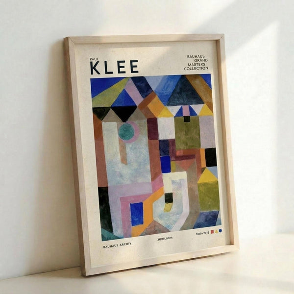 May include: Framed abstract geometric poster in light wood frame leaning against white wall. Artwork features colorful angular shapes, rectangles, triangles and circles in blues, yellows, greens, oranges and pinks with modernist Bauhaus style composition. Text reads "PAUL KLEE" at top with "BAUHAUS ARCHIV" and "1919-2019 JUBILÄUM" at bottom. Frame casts soft shadow on wall.