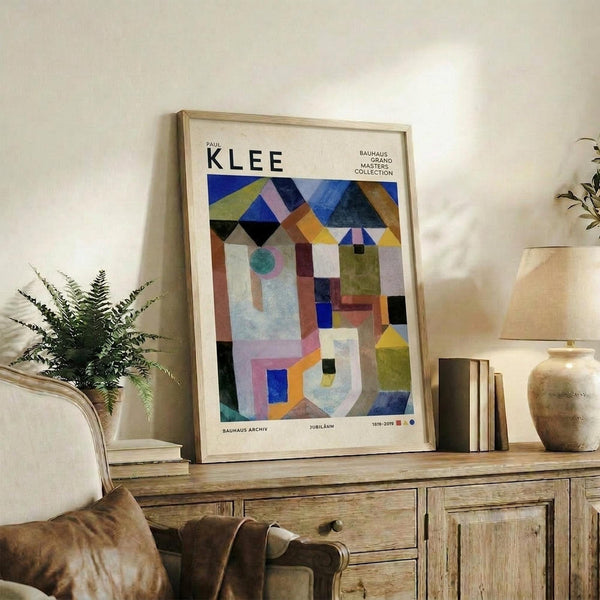 May include: Framed Paul Klee abstract art print displayed on rustic wooden sideboard in modern living room. Colorful geometric composition features overlapping shapes in blues, yellows, greens, and earth tones with Bauhaus exhibition text. Natural wood frame complements weathered furniture, fern plant, ceramic vase, and cream upholstered chair creating warm, contemporary interior styling.