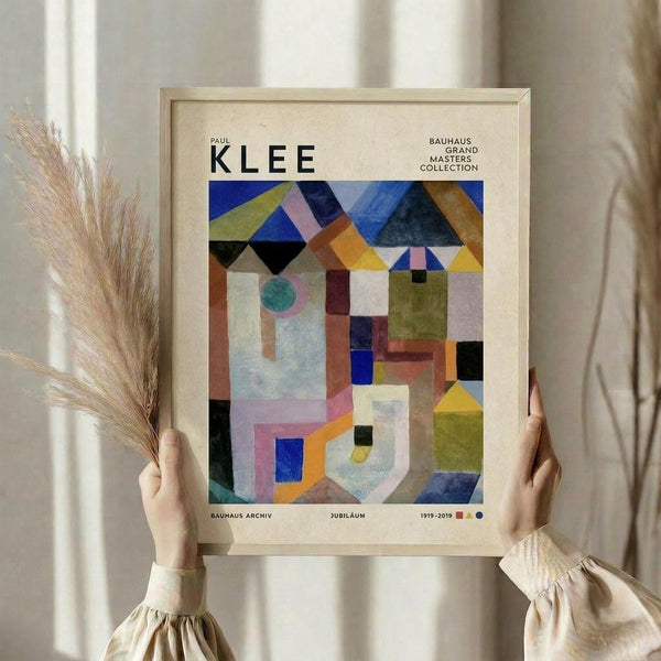 May include: Person in beige ruffled sleeves holding framed poster featuring Paul Klee's abstract geometric artwork in vibrant blues, greens, oranges, and earth tones. Light wood frame against white curtained window with dried pampas grass visible on left side.