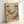 May include: Framed wall art print showing two cartoon cats relaxing on a tropical beach. One tabby cat wearing sunglasses and Hawaiian shirt sleeps in a hammock between palm trees, while an orange cat in striped shirt lounges on a beach chair. Beach scene includes ocean waves, sunset sky, cocktail glass, and small sign reading "The Art of Doing Nothing." Light wood frame against white wall corner.
