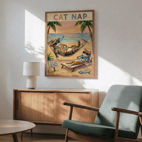 May include: Framed vintage-style poster showing two cats relaxing on a tropical beach - one sleeping in a hammock between palm trees, another lounging on a beach chair wearing sunglasses. The illustration features warm sunset colors over ocean waves with "CAT NAP" text at top and "THE ART OF DOING NOTHING" sign. The wooden-framed artwork hangs above a mid-century modern credenza with white table lamp, next to a sage green upholstered armchair in a sunlit room.