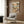 May include: Framed poster on wood-paneled wall showing cartoon cats relaxing at tropical beach - one sleeping in hammock wearing floral shirt, another on beach chair with striped shirt and cocktail, with palm trees and ocean sunset background. Modern beige armchair with geometric pillow, brass floor lamp, and dark round side table with open book in contemporary room setting.