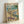 May include: Framed vintage-style poster showing two cartoon ducks doing yoga on a tropical beach. One duck in colorful floral yoga outfit performs tree pose on purple mat, while another duck in blue striped shirt sits with wine glass. Scene includes palm tree, ocean waves, wine bottles, appetizers, and beach picnic setup. Artwork has retro travel poster aesthetic with "DUCK YOGA" text in multicolored letters. Natural wood frame, displayed leaning against white wall.