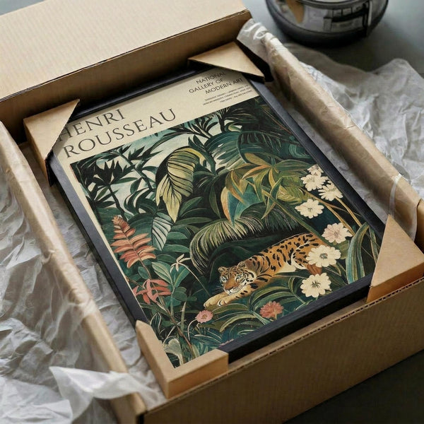 May include: Framed Henri Rousseau jungle print featuring a tiger among lush tropical foliage with pink and white flowers, displayed in black frame inside cardboard shipping box with protective tissue paper on white surface