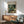 May include: Henri Rousseau jungle poster with tiger among tropical foliage displayed above mid-century modern wood credenza. Green upholstered armchair with wooden frame sits nearby. White ceramic table lamp on credenza. Warm natural lighting creates shadows on white wall. Poster shows lush botanical scene with spotted wild cat, large palm leaves, and flowering plants in rich greens and earth tones.