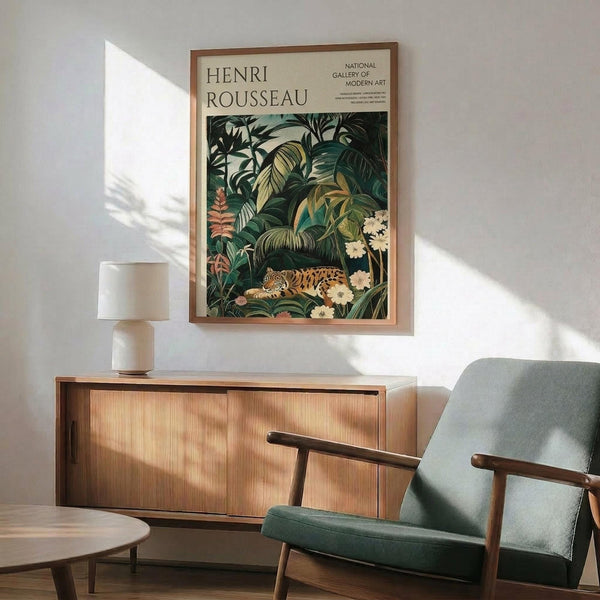 May include: Henri Rousseau jungle poster with tiger among tropical foliage displayed above mid-century modern wood credenza. Green upholstered armchair with wooden frame sits nearby. White ceramic table lamp on credenza. Warm natural lighting creates shadows on white wall. Poster shows lush botanical scene with spotted wild cat, large palm leaves, and flowering plants in rich greens and earth tones.