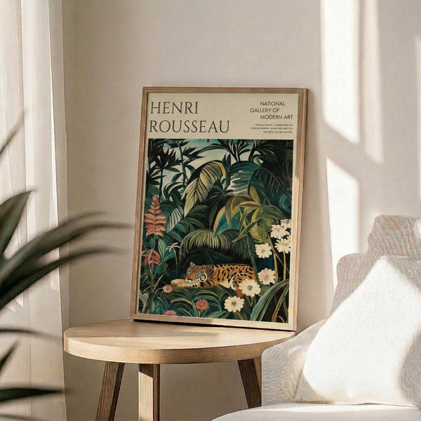 May include: Framed art print featuring Henri Rousseau's jungle scene with a leopard resting among lush green tropical foliage and white flowers, displayed on a round wooden side table next to a beige upholstered chair in a sunlit room with white walls and plant shadows.