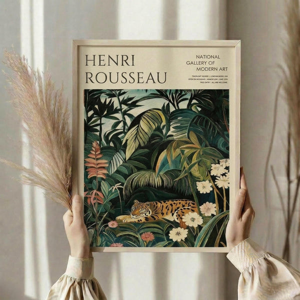 May include: Person holding framed art print featuring Henri Rousseau's jungle scene with spotted leopard resting among lush tropical foliage, palm fronds, and white flowers. Print has cream matting in light wood frame. Person wears beige ruffle-sleeve top, holds dried pampas grass stems. Soft natural lighting against neutral curtained background.