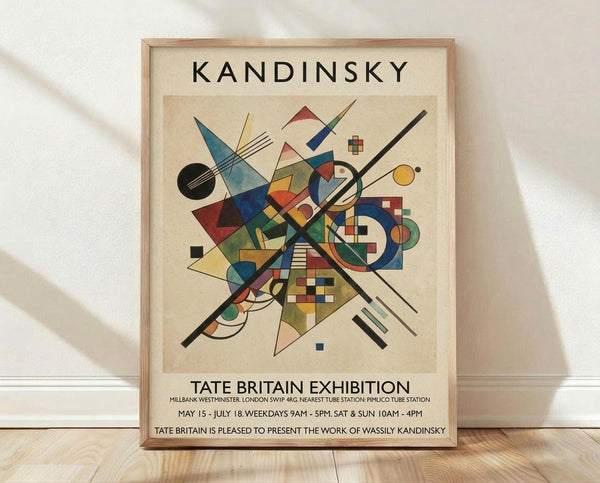 May include: Abstract geometric artwork poster in light wood frame leaning against white textured wall on wooden floor. Features colorful triangular, circular and linear shapes in blues, reds, yellows and greens on cream background. Exhibition poster format with "KANDINSKY" at top and "TATE BRITAIN EXHIBITION" text below the artwork. Soft natural lighting creates subtle shadows on wall.
