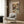 May include: Framed poster showing two white ducks in formal attire sharing a romantic dinner at an elegant table with wine, roses, and chandelier lighting, displayed on wood-paneled wall above a beige upholstered armchair with geometric throw pillow in modern living room setting with brass floor lamp and dark side table.