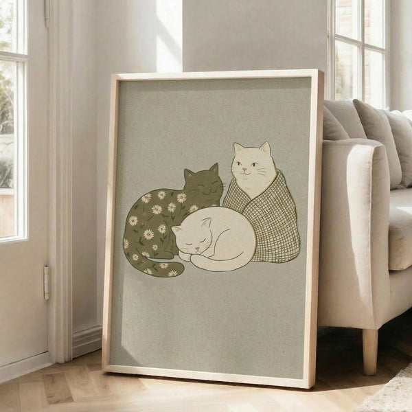 May include: Framed art print leaning against white wall next to beige armchair in bright living room. The poster features three cats in muted green and cream tones - one dark green cat with white daisy pattern, one cream cat with crosshatch texture sitting upright, and one white sleeping cat curled up. The artwork has a soft, hand-drawn illustration style on sage green background within light wood frame.