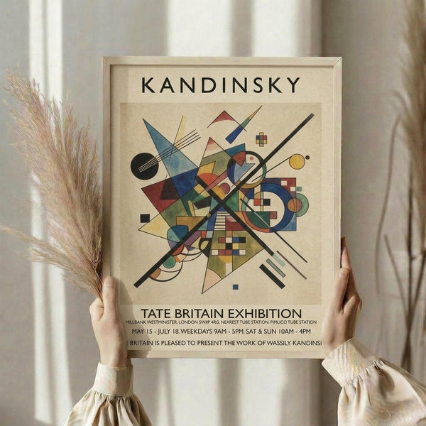 May include: Person holding framed Kandinsky abstract art print featuring geometric shapes, lines, and vibrant colors including blues, reds, yellows, and greens on cream background. Light wood frame against white curtained window with dried wheat stalks visible. Exhibition poster text shows Tate Britain details.