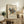 May include: Framed abstract art print displayed on rustic wooden sideboard in neutral-toned living room. Colorful geometric composition features overlapping triangles, circles, and lines in blues, reds, yellows, and greens against cream background. Exhibition poster format with black text below artwork. Scene includes fern plant, ceramic vase, stacked books, and beige upholstered furniture creating warm, modern home decor setting.