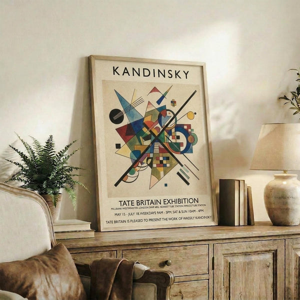 May include: Framed abstract art print displayed on rustic wooden sideboard in neutral-toned living room. Colorful geometric composition features overlapping triangles, circles, and lines in blues, reds, yellows, and greens against cream background. Exhibition poster format with black text below artwork. Scene includes fern plant, ceramic vase, stacked books, and beige upholstered furniture creating warm, modern home decor setting.