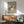 May include: Framed poster featuring three cats in soft green and cream tones on sage background, displayed above mid-century modern wooden credenza with white ceramic lamp. Green upholstered armchair with wooden frame sits nearby in bright, naturally-lit room with white walls and warm wood furnishings.
