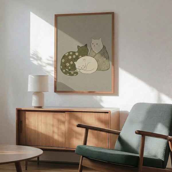 May include: Framed poster featuring three cats in soft green and cream tones on sage background, displayed above mid-century modern wooden credenza with white ceramic lamp. Green upholstered armchair with wooden frame sits nearby in bright, naturally-lit room with white walls and warm wood furnishings.