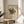 May include: Framed Kandinsky abstract art poster with geometric shapes, lines, and vibrant colors in red, blue, yellow, and green, displayed on a round wooden stool next to a beige upholstered chair. Natural sunlight streams across the white wall, with a potted plant visible in the corner. The poster features exhibition text at the bottom and is presented in a thin wooden frame.