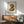 May include: Framed abstract art print featuring two balanced oval stones in earthy brown and sage green tones against cream background, displayed above mid-century modern walnut credenza with ceramic table lamp, next to teal upholstered armchair with wooden arms in sunlit room with white walls.