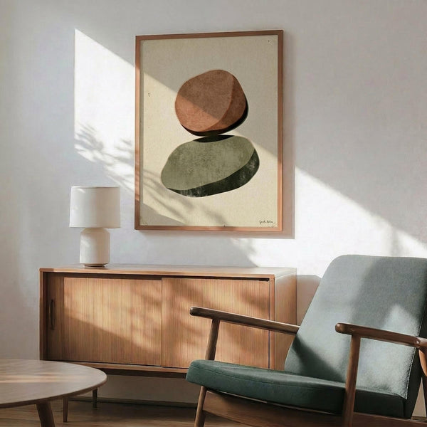 May include: Framed abstract art print featuring two balanced oval stones in earthy brown and sage green tones against cream background, displayed above mid-century modern walnut credenza with ceramic table lamp, next to teal upholstered armchair with wooden arms in sunlit room with white walls.