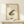 May include: Framed vintage-style artwork featuring a spotted brown and black frog with Japanese text and red stamps, displayed on a light wooden shelf alongside a small white wrapped gift with evergreen sprig. The beige textured paper artwork is housed in a natural wood frame against a cream wall, creating a minimalist aesthetic.