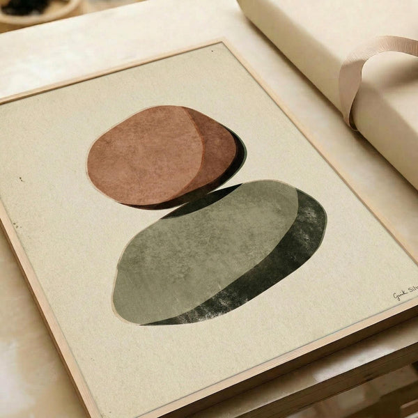 May include: Abstract minimalist art print featuring two overlapping oval shapes with textured surfaces - one in warm terracotta brown tones above, one in sage green below, both casting subtle shadows on cream background. The artwork is displayed in a light wood frame on white paper, photographed on a neutral surface with soft natural lighting and ribbon visible nearby.