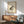 May include: Framed vintage-style frog artwork hanging on white wall above wooden sideboard with table lamp, next to mid-century modern teal upholstered armchair. The print features a spotted brown and black frog on aged cream paper with Asian calligraphy and red seal stamps in upper left corner. Natural sunlight creates dramatic shadows across the minimal, warm-toned interior scene.