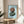 May include: Framed art print featuring two balanced stones in brown and sage green tones on a light blue background, displayed on a round wooden side table next to a beige upholstered chair. Natural sunlight streams across the white wall, creating soft shadows. A plant's leaves are partially visible in the corner, and the wooden frame complements the warm, minimalist Scandinavian-style interior.