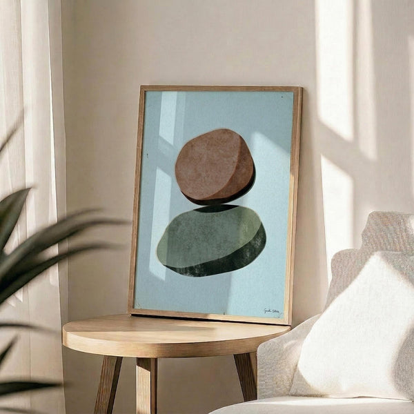 May include: Framed art print featuring two balanced stones in brown and sage green tones on a light blue background, displayed on a round wooden side table next to a beige upholstered chair. Natural sunlight streams across the white wall, creating soft shadows. A plant's leaves are partially visible in the corner, and the wooden frame complements the warm, minimalist Scandinavian-style interior.