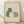 May include: Framed artwork displaying three stylized cats in muted earth tones - one green cat with white daisy pattern, one cream-colored cat, and one with green checkered pattern. The cats are arranged in a cozy group against a beige textured background. Natural wood frame sits on light wooden flooring against white wall with soft shadows.