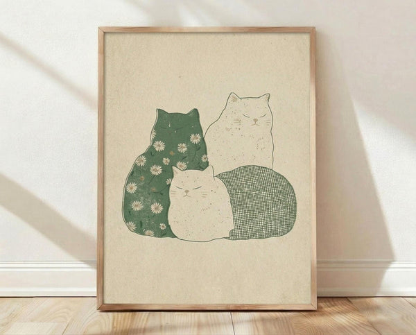 May include: Framed artwork displaying three stylized cats in muted earth tones - one green cat with white daisy pattern, one cream-colored cat, and one with green checkered pattern. The cats are arranged in a cozy group against a beige textured background. Natural wood frame sits on light wooden flooring against white wall with soft shadows.