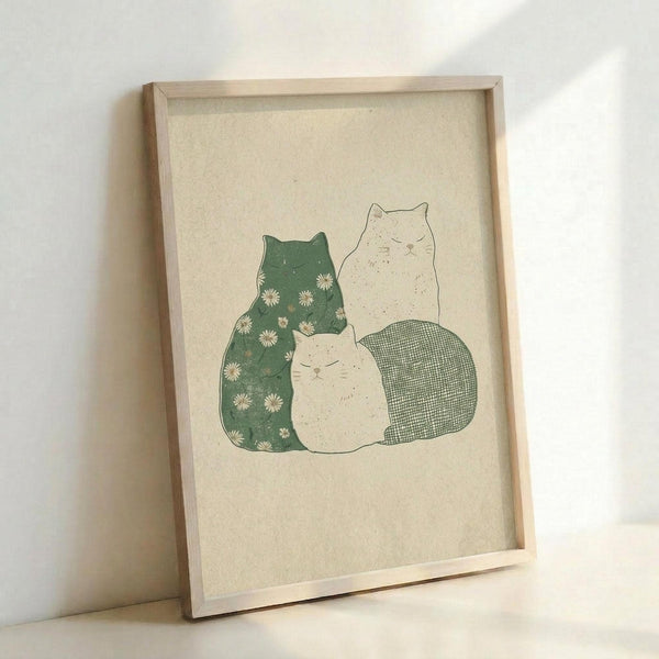 May include: Framed art print showing three stylized cats in soft green and cream colors on textured beige paper. The left cat has a green body decorated with small white daisy flowers, while two cream-colored cats sit beside it, one with a green crosshatch pattern on its lower body. The illustration has a gentle, hand-drawn aesthetic displayed in a light wood frame against a white wall.