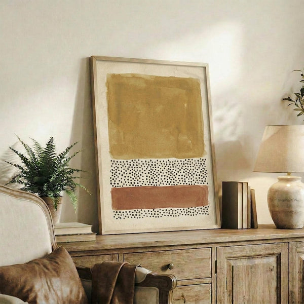 May include: Modern abstract art print in light wood frame displaying warm ochre rectangle, dotted pattern section, and terracotta brushstroke on cream background. Styled on rustic wooden sideboard with leather chair, fern plant, ceramic vase, and books in neutral-toned living room setting.