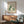 May include: Framed cat artwork showing three cats in green daisy pattern, cream, and teal colors displayed above a wooden sideboard with ceramic lamp in modern living room with mint green upholstered chair and natural sunlight shadows on white walls.