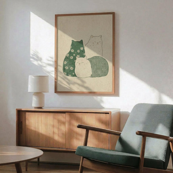 May include: Framed cat artwork showing three cats in green daisy pattern, cream, and teal colors displayed above a wooden sideboard with ceramic lamp in modern living room with mint green upholstered chair and natural sunlight shadows on white walls.