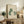 May include: Framed artwork showing three cats in soft green and cream colors, with one cat featuring a white daisy pattern, displayed on a rustic wooden sideboard alongside books, a ceramic vase, and a fern plant in a cozy living room setting with neutral tones and natural lighting.